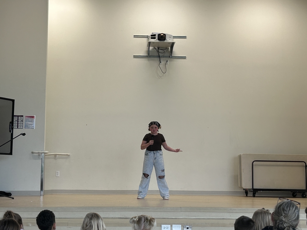  A student performer stands in the center of a wooden stage, holding a microphone and gesturing with one hand while performing. They are wearing a dark t-shirt, light-colored distressed jeans with holes at the knees, and a patterned bandana on their head. The background is a plain, high-ceilinged wall with a projector mounted above, and the tops of the audience's heads are visible in the foreground.