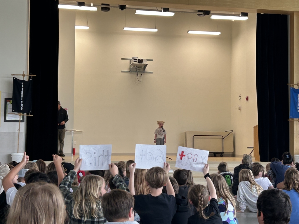  A young student stands center stage at a microphone during a school talent show. In the foreground, several students in the audience hold up handmade signs to support the performer. The stage is framed by black curtains, with a blue house banner visible on the right and a black one on the left. A projector is mounted on the back wall above the performer, and an adult stands partially visible in the wings.