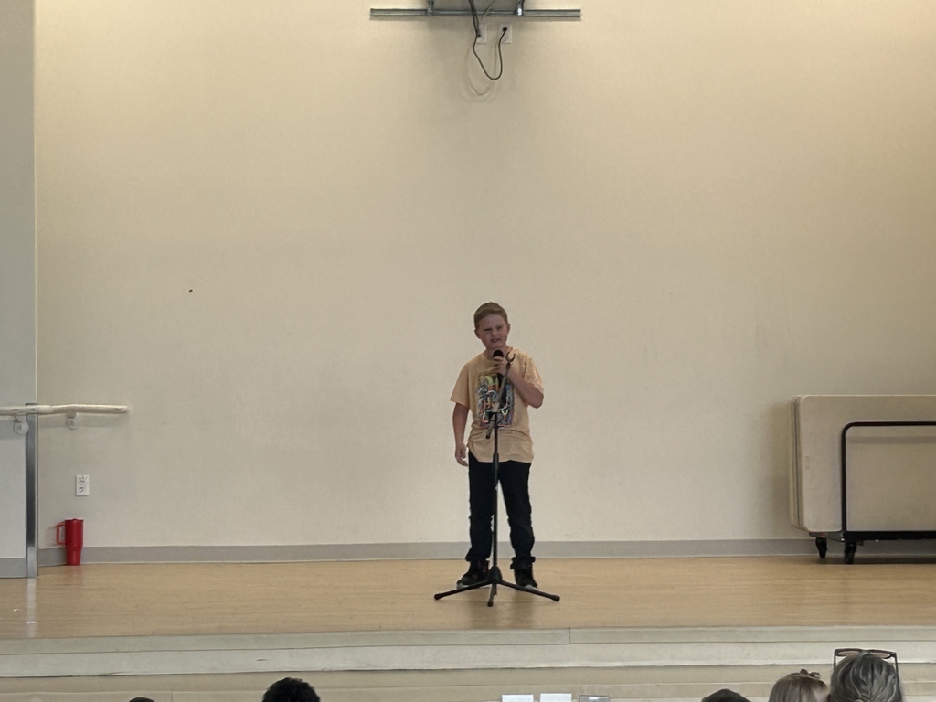 An elementary-aged boy stands alone on a wooden stage, holding a microphone to his mouth and performing for an audience. He is wearing a tan graphic t-shirt, dark pants, and sneakers. The background is a large, plain cream-colored wall, and the top of the audience's heads are visible at the bottom of the frame.