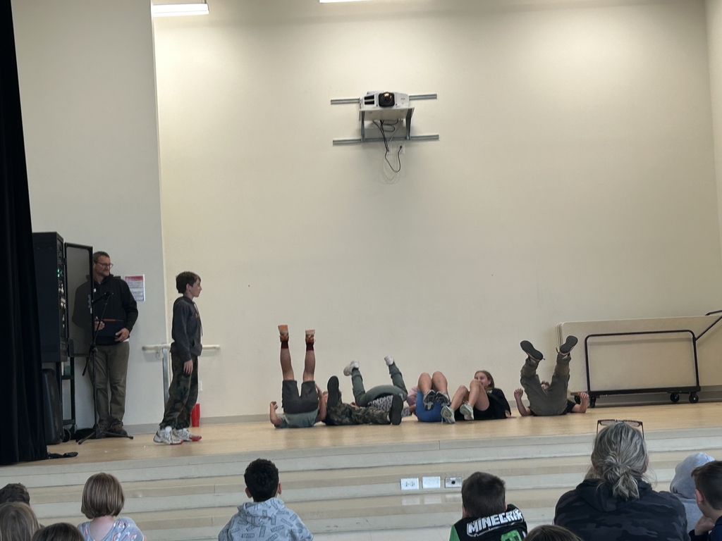  A group of students performs a comedic skit on a wooden stage during a school talent show. Five students are lying on their backs in a row with their legs kicked up in the air, while another student stands to the left watching them. An adult stands near sound equipment in the wings, and the back of an audience is visible at the bottom of the frame under a mounted ceiling projector.