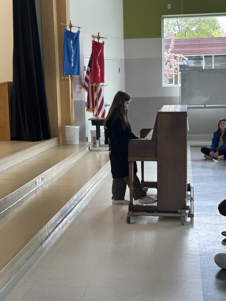 A student with long hair stands at an upright wooden piano on wheels, performing in a school multipurpose room. They are wearing a black hoodie and brown pants. In the background, a blue house banner, a red house banner, and the American flag are displayed next to the stage. Another student sits on the floor in the distance near a whiteboard and a large window.