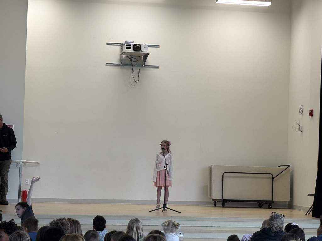 A young girl stands center stage at a microphone, performing for a large audience during a school talent show. She is wearing a white cardigan over a pink dress with a large pink bow in her hair. An adult stands partially visible on the far left, and a projector is mounted on the wall above the performer. The tops of the audience members' heads are visible in the foreground, with one student raising a hand.