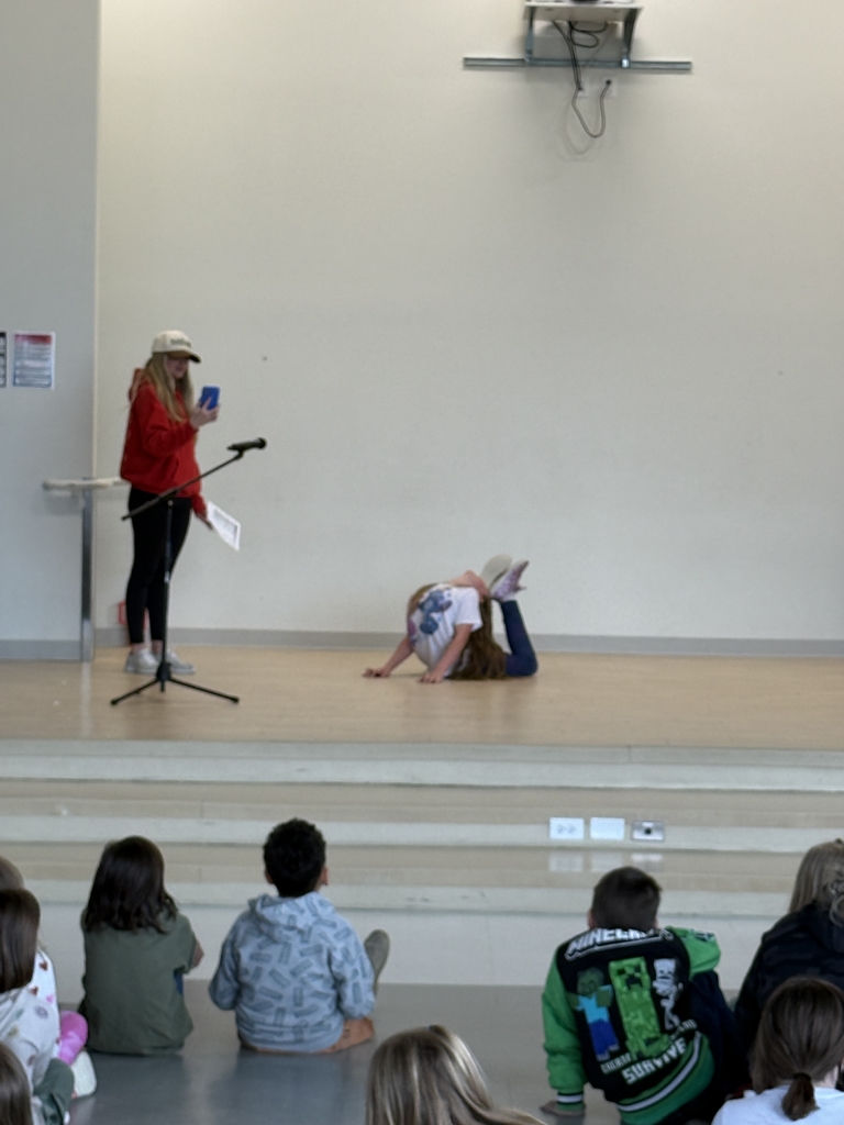  A student performer on a wooden stage demonstrates extreme flexibility by performing a deep backbend while lying on their stomach. To the left, an older student in a red hoodie and baseball cap stands by a microphone, holding a phone. In the foreground, several elementary students sit on the floor watching the performance, including one student wearing a green Minecraft-themed jacket.