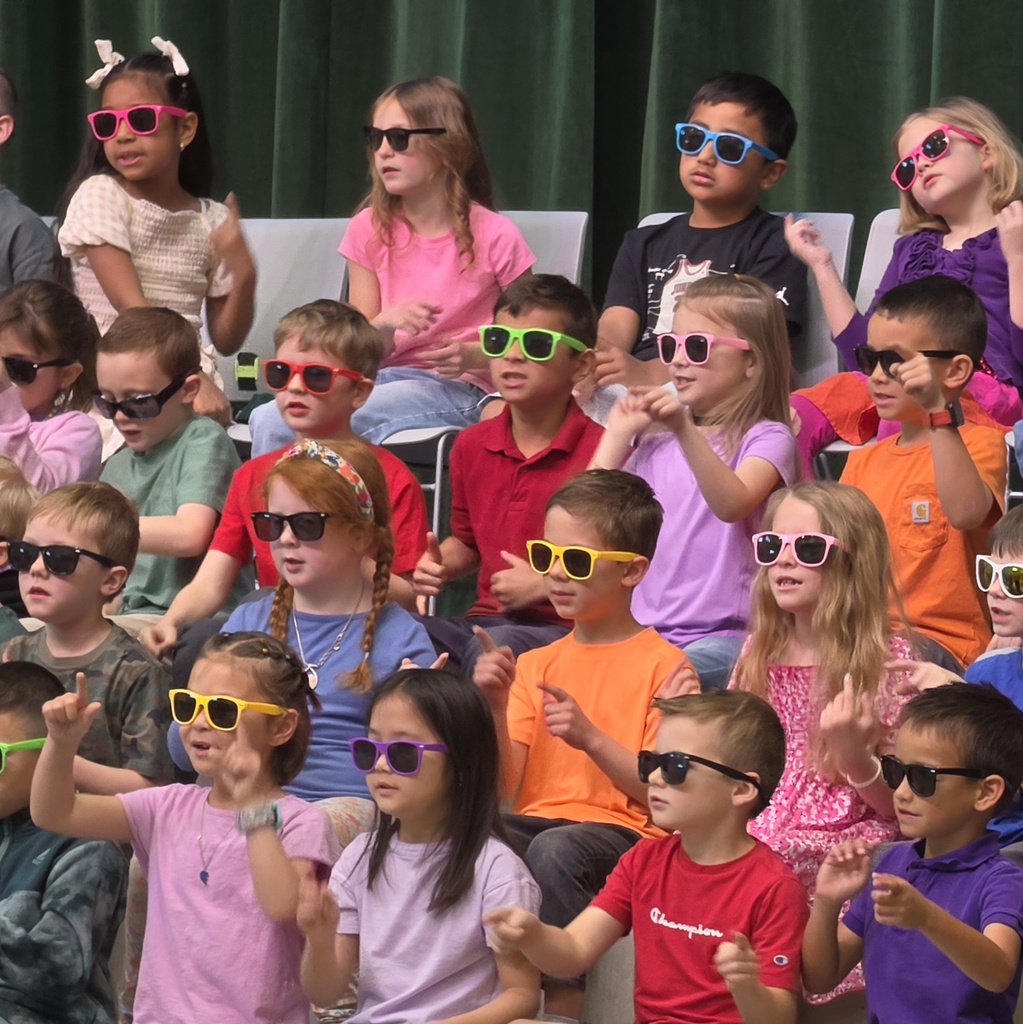 Students in bright sunglasses perform together on risers during the first-grade Spring Sing.