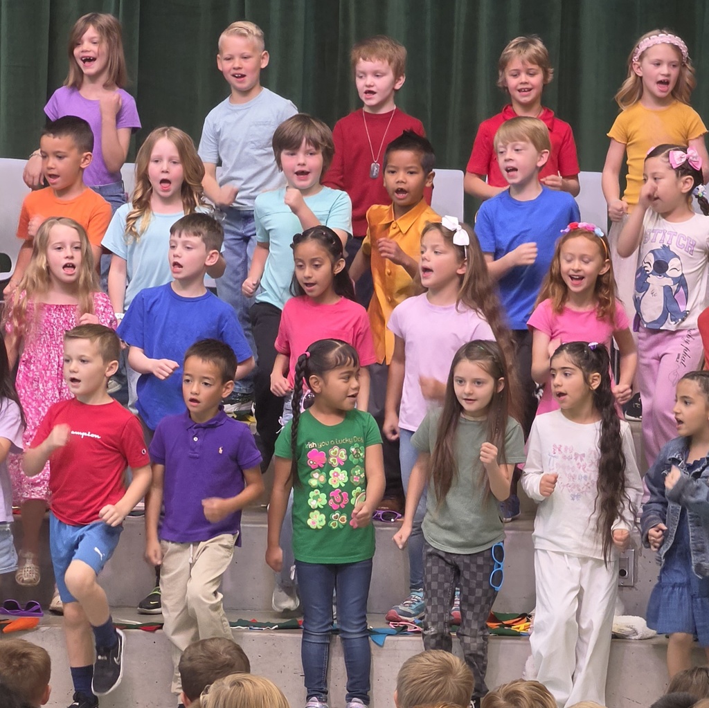 A group of first-grade students sings on risers, using small arm motions as part of their Spring Sing performance.