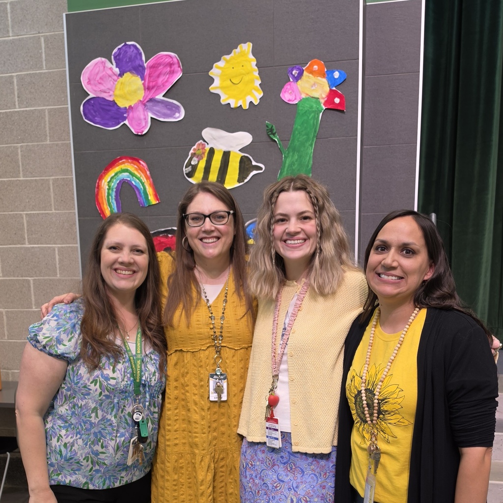 Four first-grade teachers smile together in front of colorful student-made spring artwork after the Spring Sing performance.