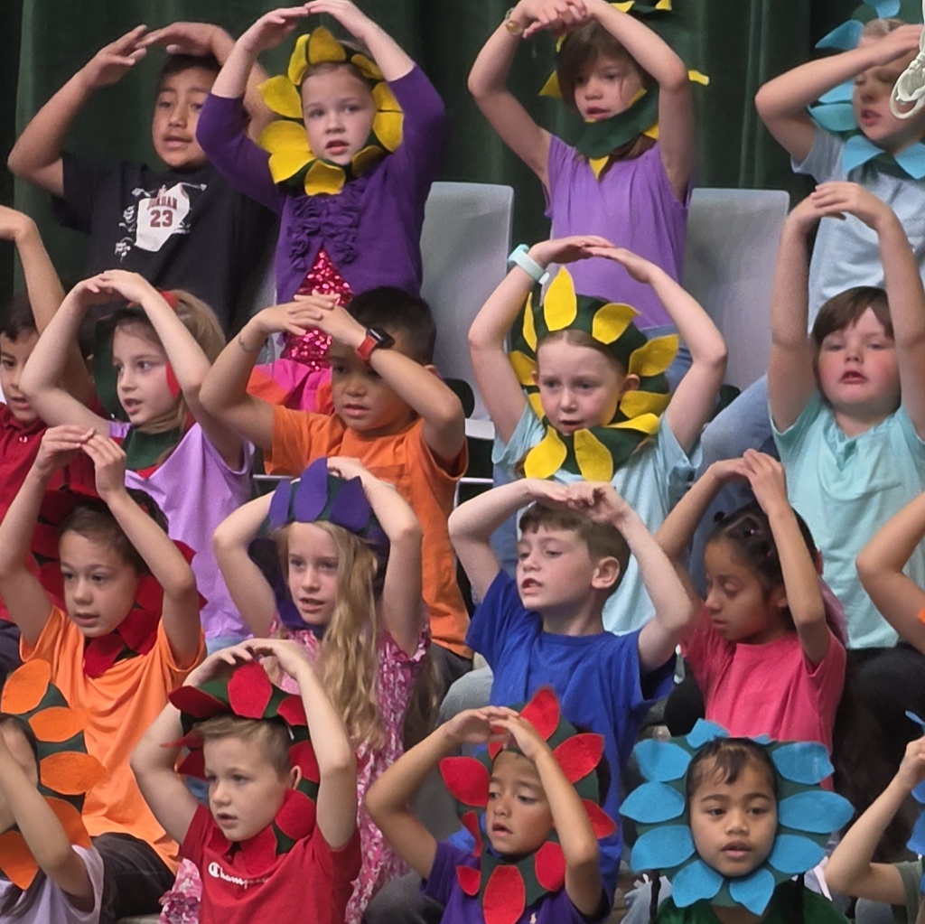Students wearing flower headpieces raise their arms in rounded shapes while performing spring-themed motions on stage.