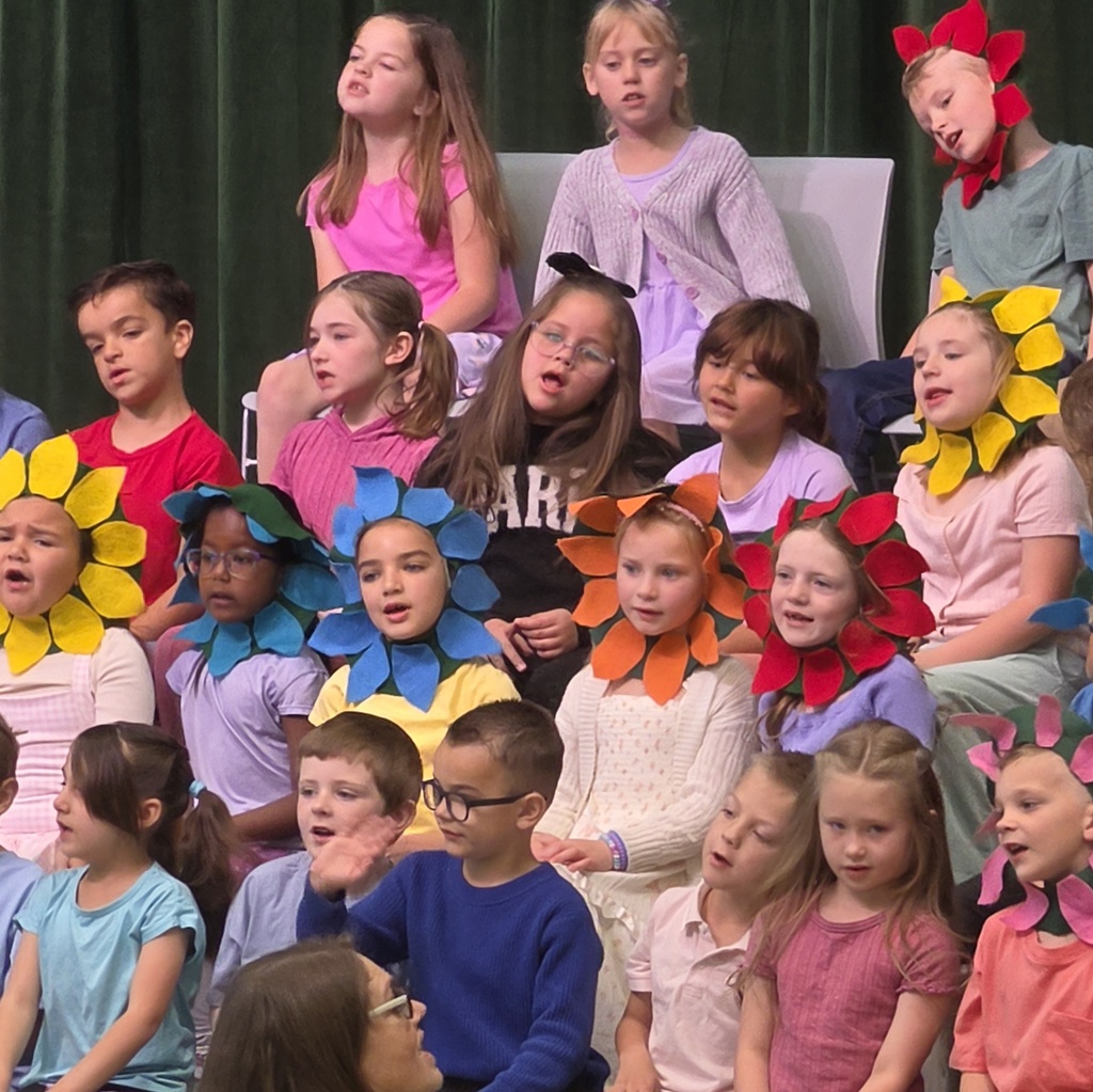 First-grade students sing on risers, with several students wearing colorful flower headpieces for the spring-themed performance.
