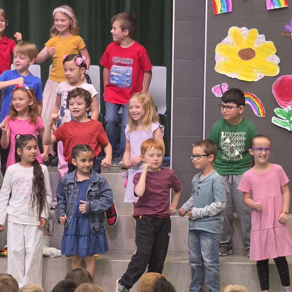 First-grade students smile and perform on stage beside colorful student-made spring artwork, including flowers and rainbows.