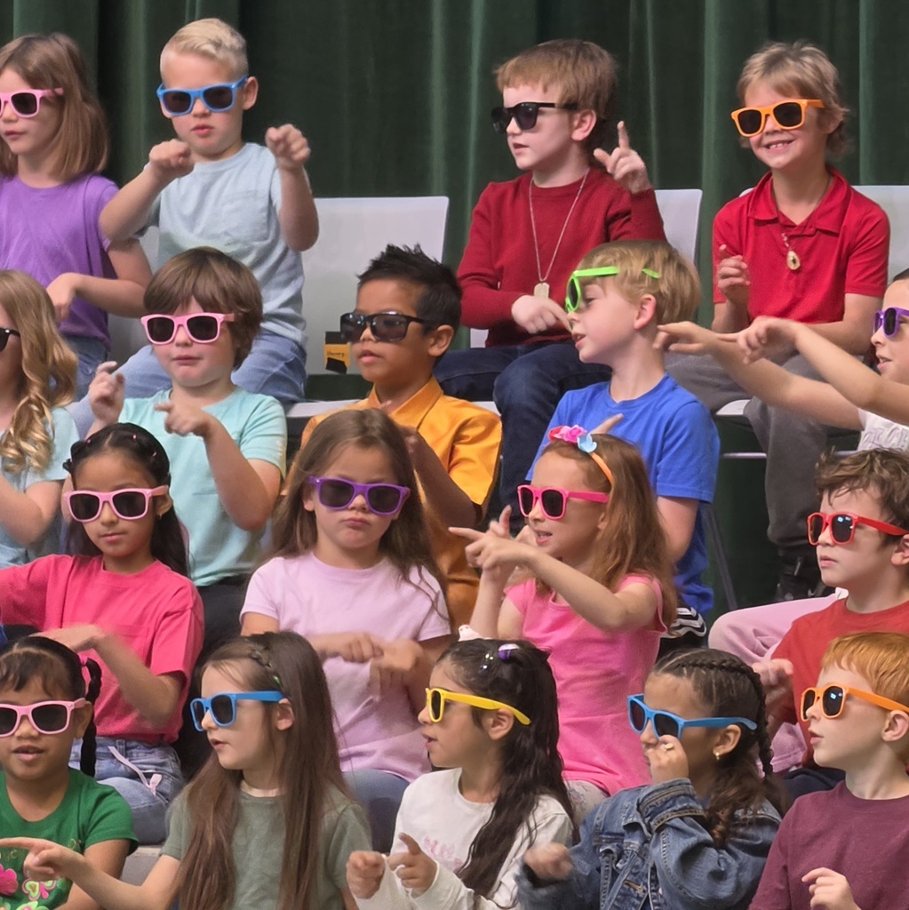 First-grade students wear colorful sunglasses while singing and doing hand motions during their Spring Sing performance.