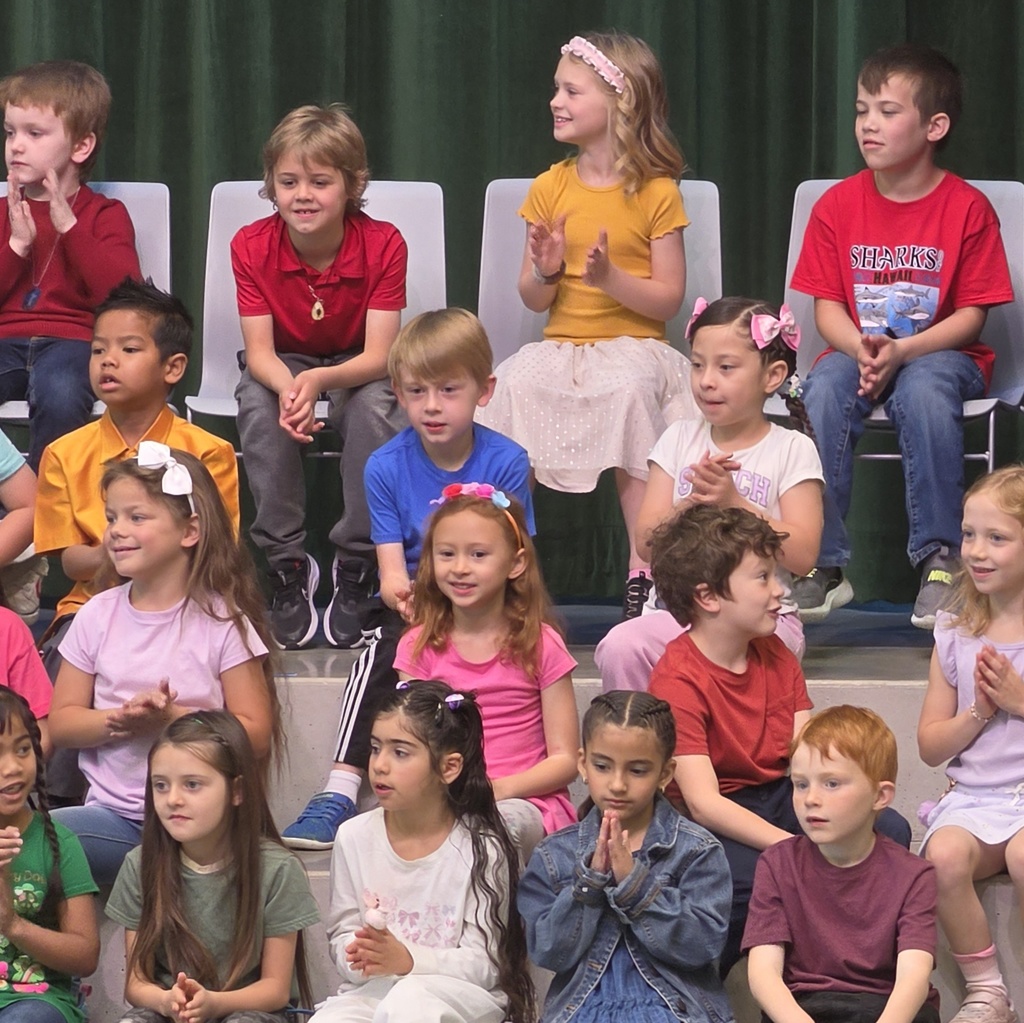 First-grade students sit and clap on risers in front of a green curtain during their Spring Sing performance.