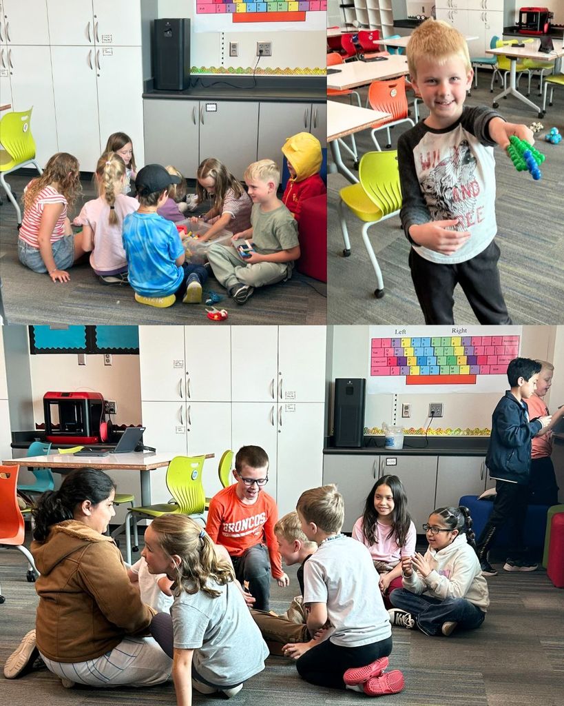 Elementary students sit in small groups on the classroom floor building and playing with materials, while another student stands smiling and showing a colorful creation in front of desks and bright chairs.