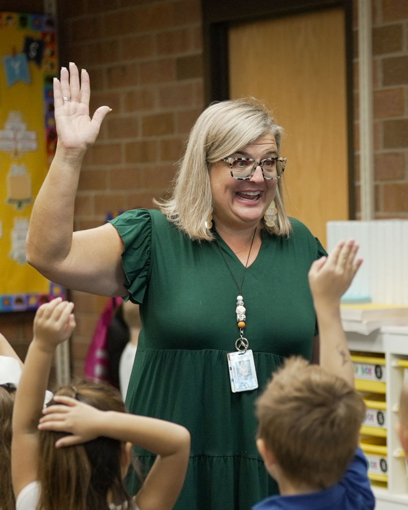 A principal raising her hand happily with students surrounding her doing the same. 