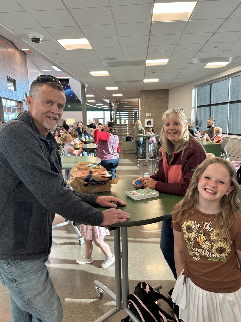 blonde girl with brown shirt smiling at camera with grown man and lady with books in the background