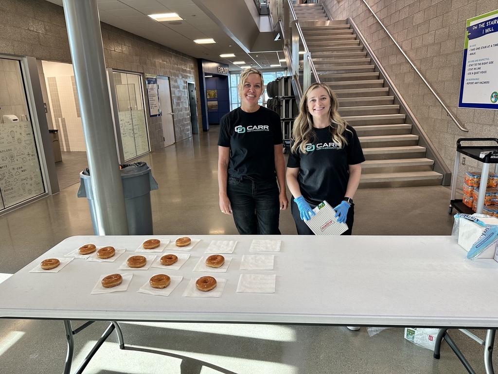 two ladies in black shirts standing behind a table with donuts on it