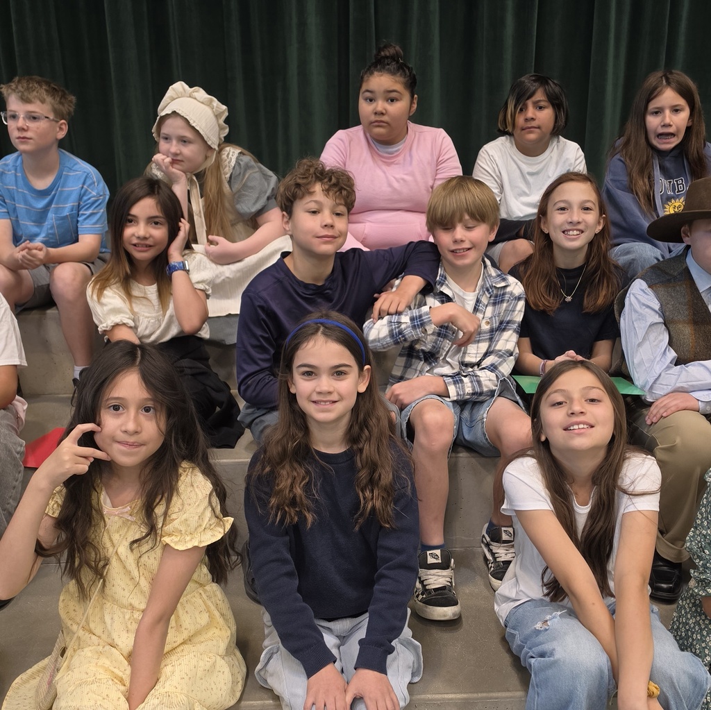 Fourth grade students sit together on risers before their program. Several students smile at the camera, while others wear pioneer-inspired costumes and casual school clothes.