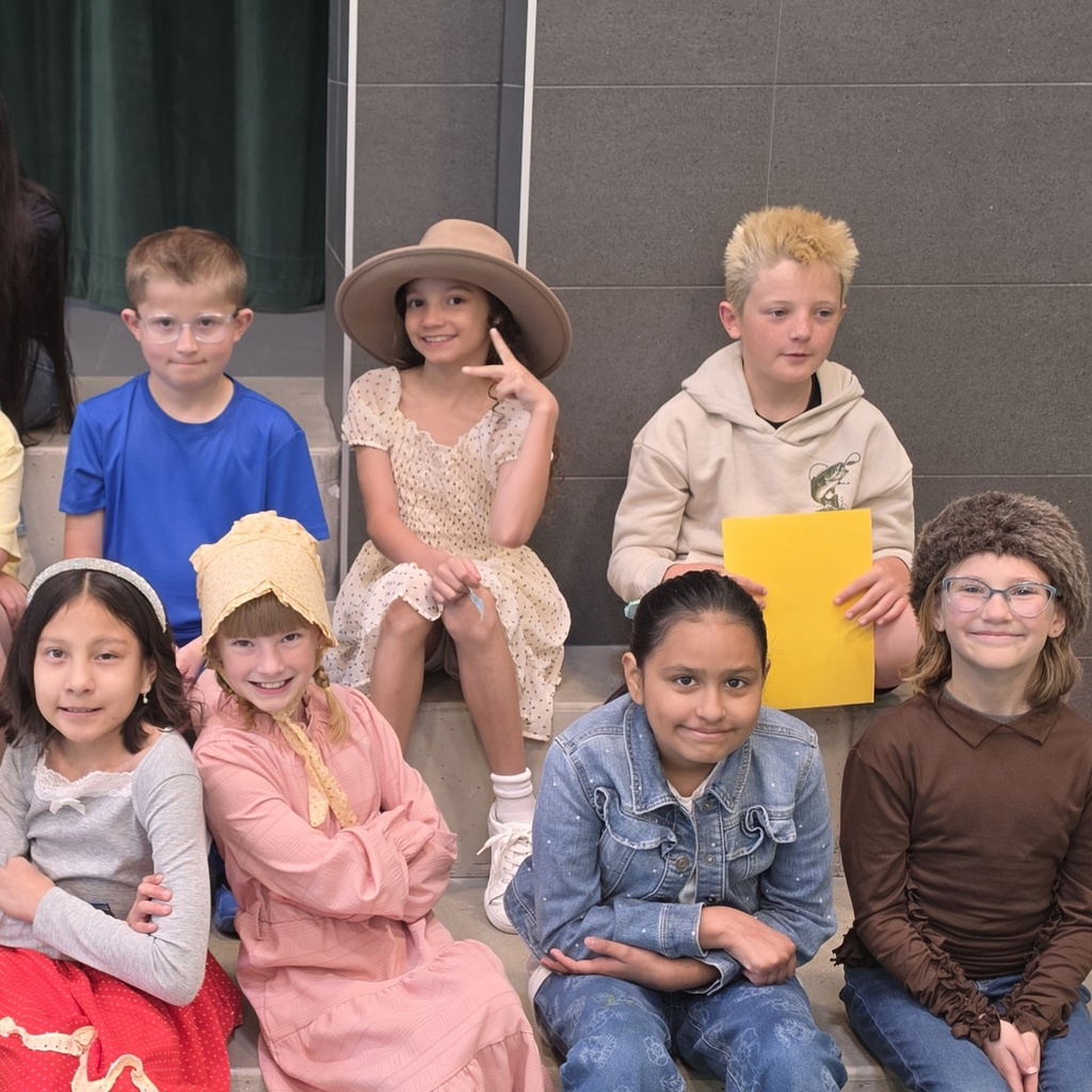 Fourth grade students sit on risers before their program. Some students smile at the camera while others hold papers or wear costume pieces connected to the Utah history theme.