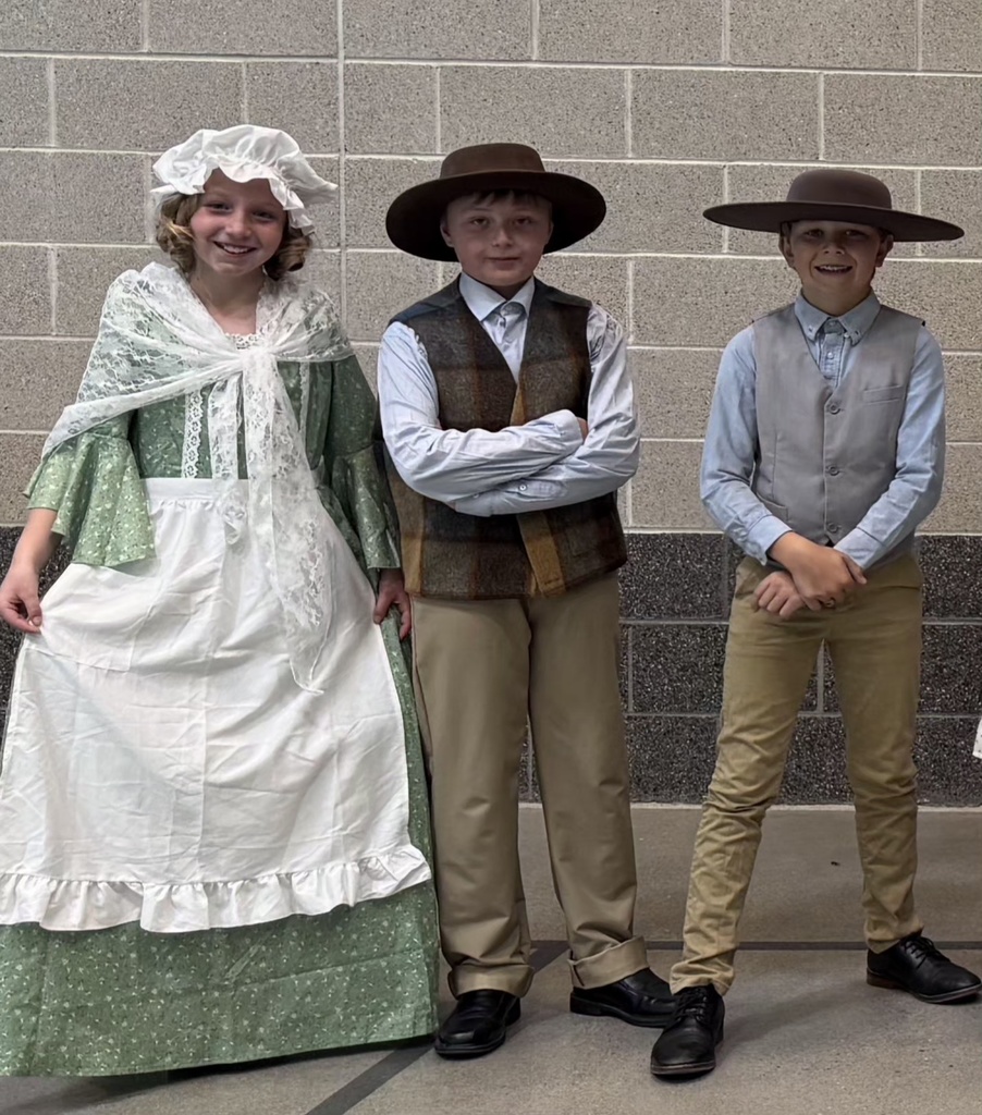 Students in pioneer-style costumes pose together in front of a gym wall after the Utah history program. Costumes include bonnets, wide-brimmed hats, vests, suspenders, and dresses.