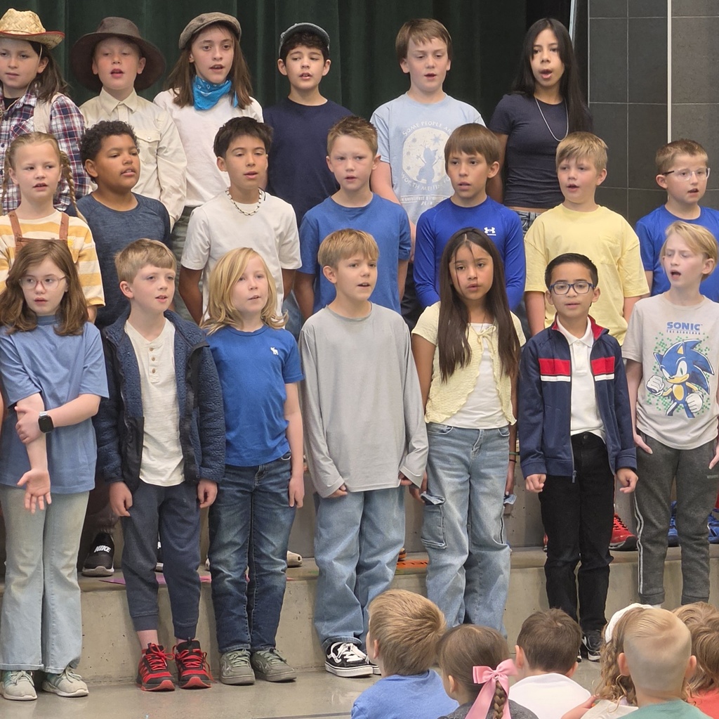 A group of fourth grade students stands on risers singing during their Utah history program. Some students wear pioneer-inspired clothing while others are dressed in everyday school clothes.
