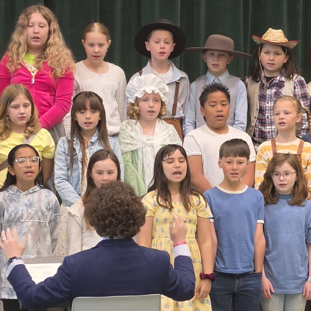 Fourth grade students sing on risers while a teacher directs them from the front. Several students wear pioneer-style hats, bonnets, and dresses for the Utah history performance.