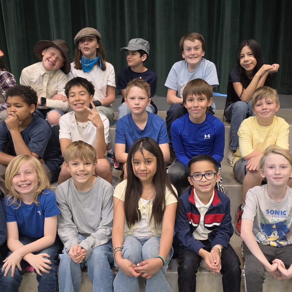 Fourth grade students sit together on risers and smile for a group photo after their Utah history program. Some students wear costume pieces such as hats, while others are dressed casually.