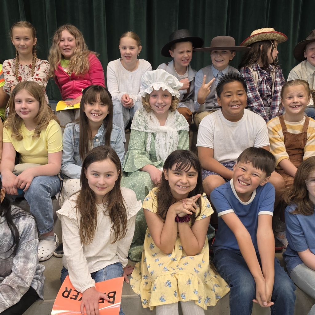 Fourth grade students sit together on risers and smile for a group photo after their Utah history program. Some students wear costume pieces such as hats, while others are dressed casually.