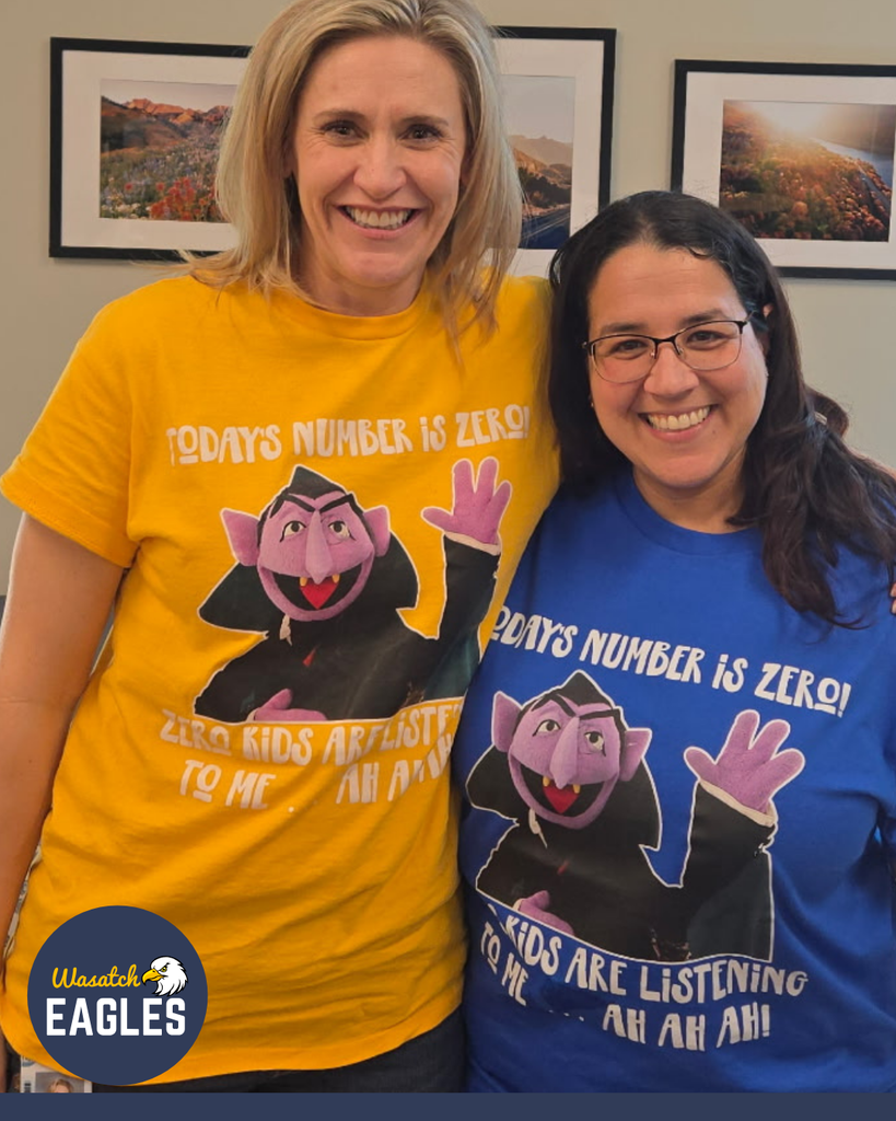 Two adults stand close together indoors, facing the camera. One wears a yellow T‑shirt and the other wears a blue T‑shirt. Both shirts feature a cartoon character and the phrases “Today’s number is zero” and “Zero kids are listening.” Framed landscape photographs hang on the wall behind them, and part of a school logo appears in the lower corner of the image.