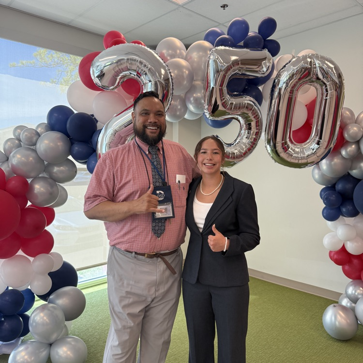 Two people smiling in front of a red, white, and blue balloon arch with silver “250” balloons. One person is wearing a pink shirt and tie, and the other is wearing a dark suit and giving a thumbs up.