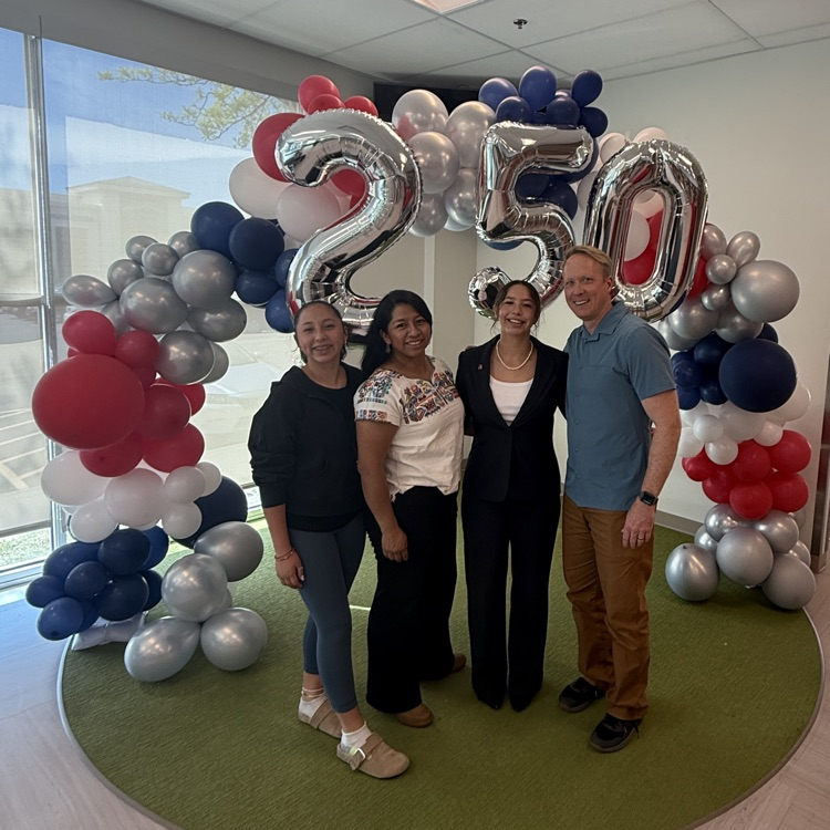 Four people posing in front of a red, white, blue, and silver balloon arch with silver “250” balloons indoors.