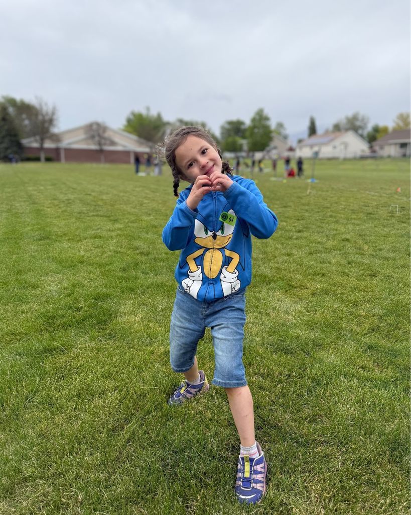 A smiling student stands on a grassy field during the Unified Yard Game Olympics, making a heart shape with their hands. The student is wearing a blue Sonic hoodie, jean shorts, and sneakers while other students and game stations can be seen blurred in the background under a cloudy sky.