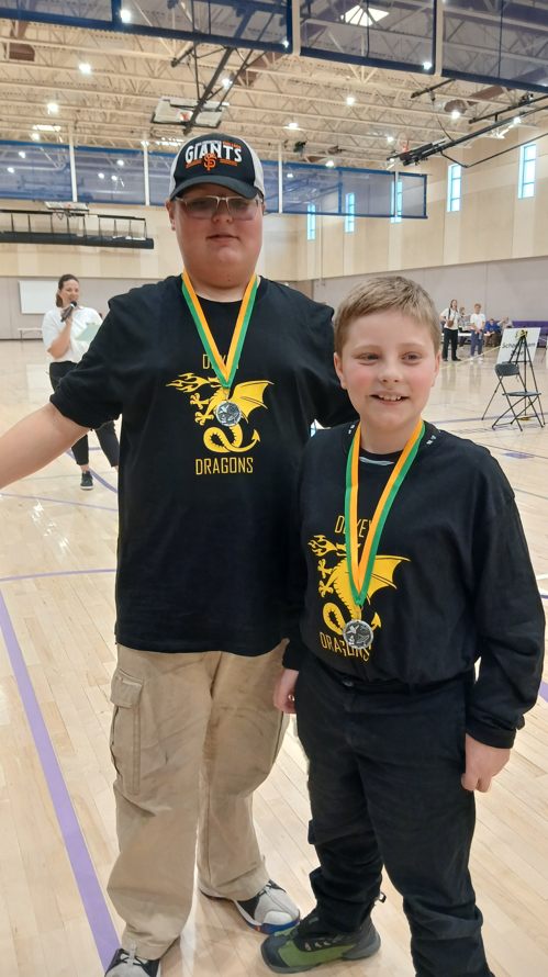 Two boys from the Doxey Elementary team stand proudly in the center of the gymnasium wearing their silver 2nd place medals with green and yellow ribbons.