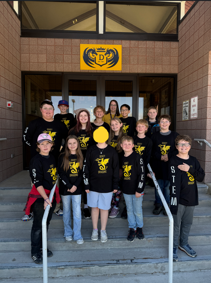 A group photo of the Doxey Elementary STEM Olympiad team wearing matching black long-sleeve shirts with a yellow dragon logo. They are posed together on the concrete steps in front of the school’s main entrance under a yellow Doxey Dragons sign.