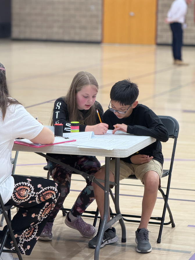Two Doxey Elementary students sit together at a white folding table in a gym, working collaboratively on a "Data Detectives" paper-based challenge. A judge with a clipboard sits at the table with them to monitor their progress.