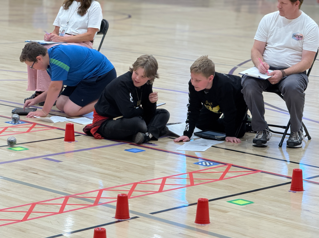 Two students from the Doxey team are kneeling on a gymnasium floor, intently focused on a Sphero robotics course. They are using a tablet to navigate the small robotic balls through a path marked by red tape and plastic cups while judges watch from behind.