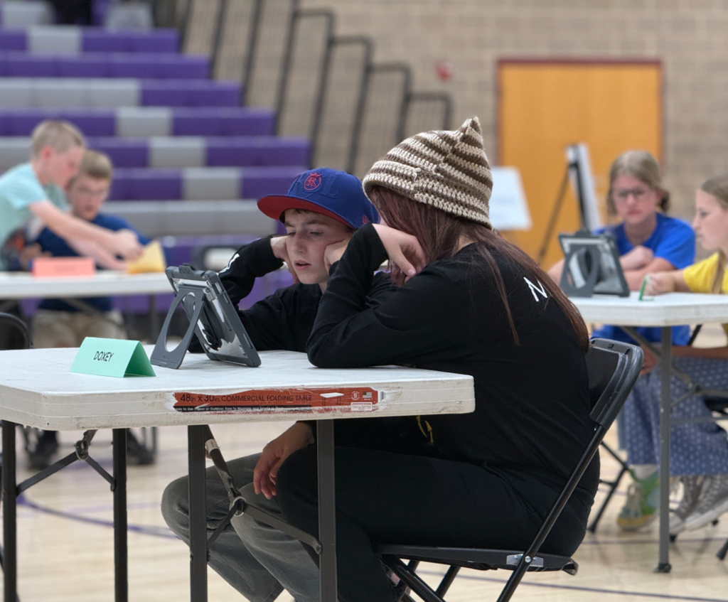 Two students in Doxey Dragons shirts are seated at a competition table, leaning in close to study a tablet together. In the background, other teams are visible at their own stations in the gymnasium.