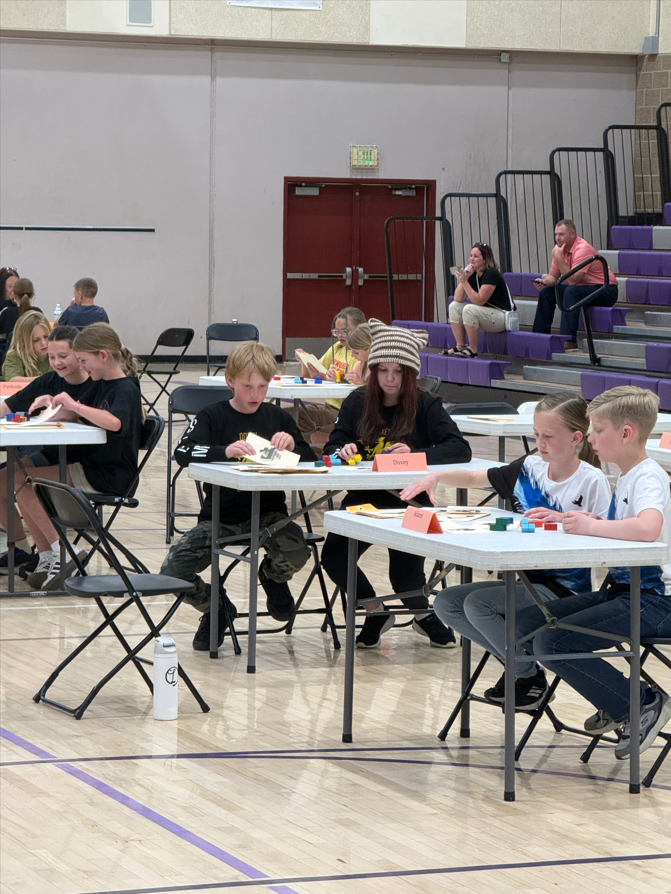 Several Doxey students are seated at tables in a gymnasium, focused on a "Soma Cubes" event. They are manipulating small, colorful blocks while competing alongside students from other schools.