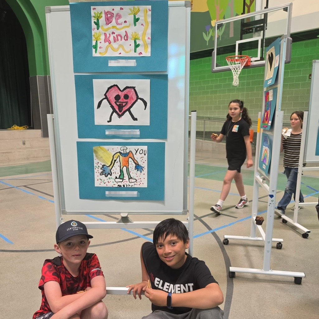 Bountiful Elementary students sit beside a display of Kind Kids Club artwork celebrating kindness, friendship, and positivity.
