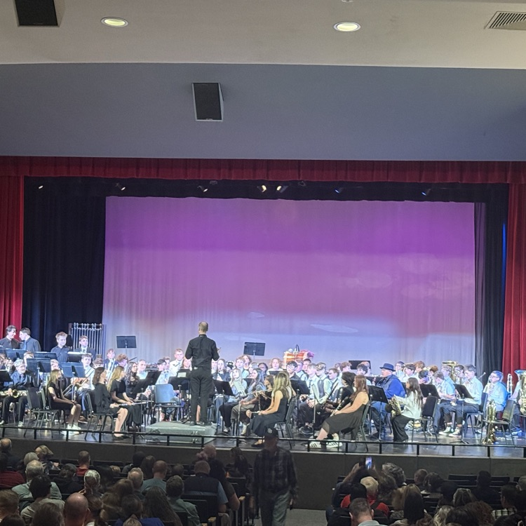 A school band performs on stage in an auditorium while a conductor leads from the center. Audience members sit in front of the stage