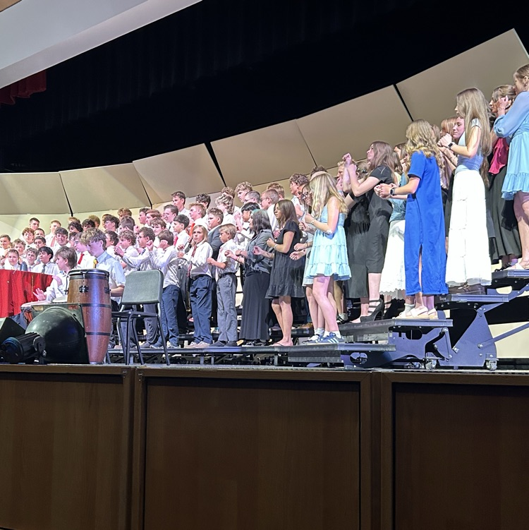 A large school choir stands on risers during a concert performance on stage. Students are dressed in concert attire, and percussion instruments are visible near the front of the stage