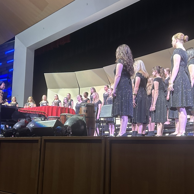 A group of students in black concert attire stands on risers during a choir performance on stage. A piano, percussion instruments, and stage lighting are visible near the front,