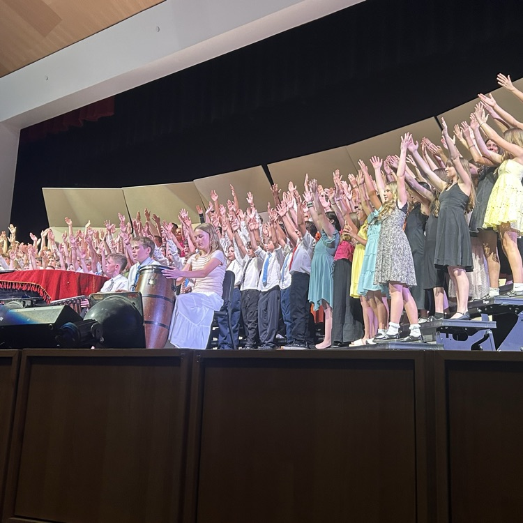 A large group of students performs on stage during a school concert with their arms raised in the air. Students stand on risers in concert attire, with percussion instruments and stage equipment visible at the front.