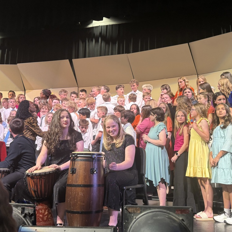 A large group of students performs on stage during a school concert with their arms raised in the air. Students stand on risers in concert attire, with percussion instruments and stage equipment visible at the front.