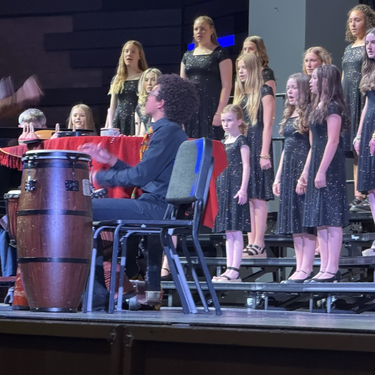 A student plays hand drums at the front of a school choir performance while singers in matching black concert attire stand on risers behind him. Stage lighting and percussion instruments are visible during the performance. 