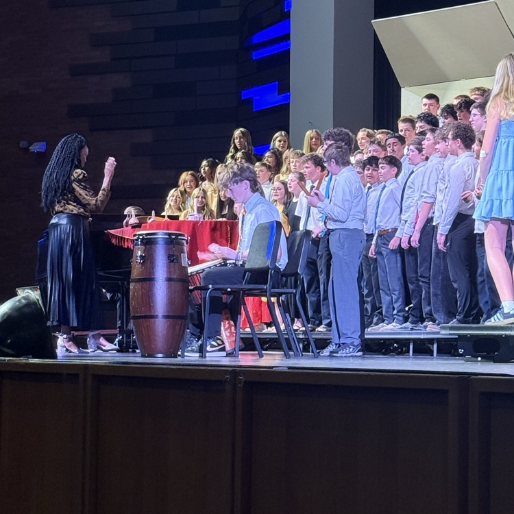 A choir director leads students during a school concert while a student plays percussion at the front of the stage. Choir members stand on risers in concert attire under blue stage lighting. 