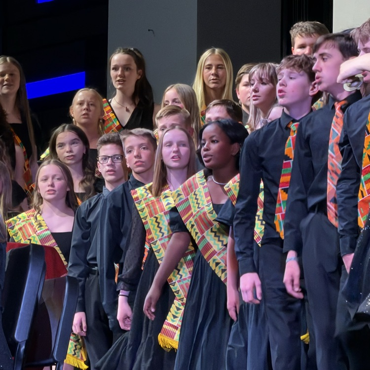 Students sing during a school choir performance while wearing black concert outfits with colorful patterned scarves and ties. Choir members stand closely together on risers under stage lighting. 
