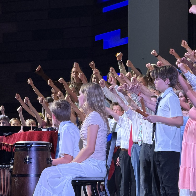 Students perform on stage during a school concert with many raising their fists in the air as part of the performance. Percussion instruments and seated student musicians are visible at the front of the stage under blue stage lighting.