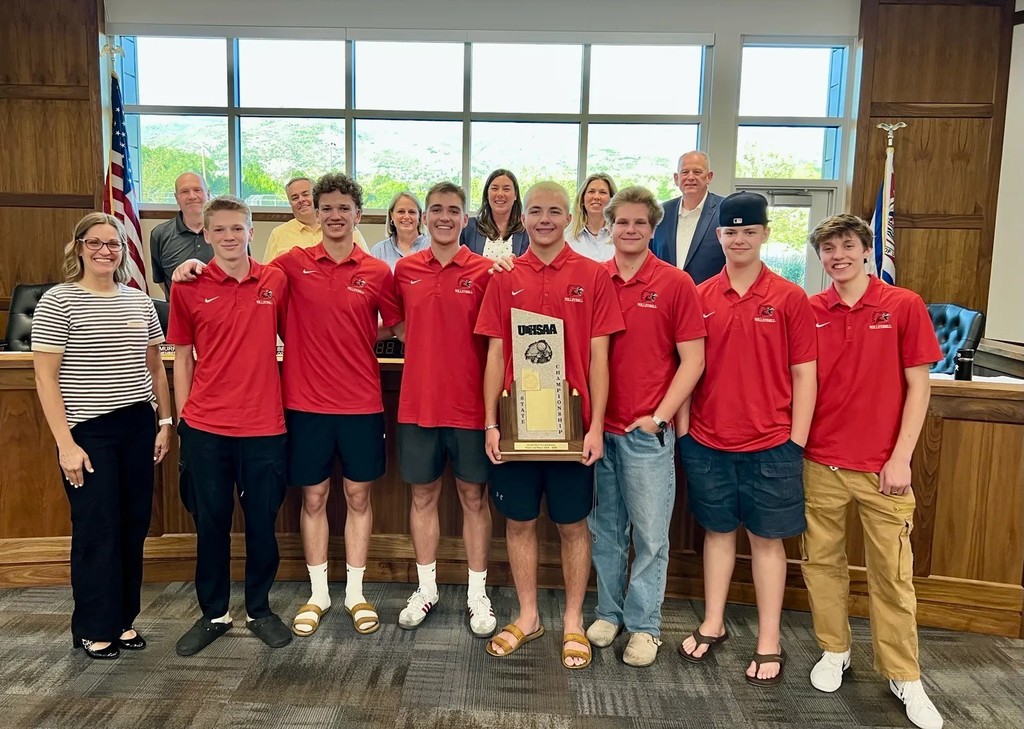 Volleyball team standing at the front of the City Council room with city councilmen and women and their trophy. 