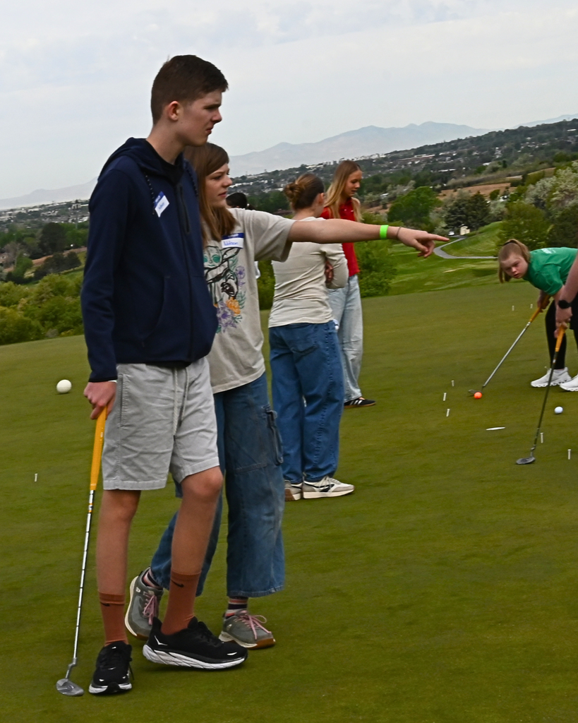 A student standing next to another student golfing and pointing to the right.
