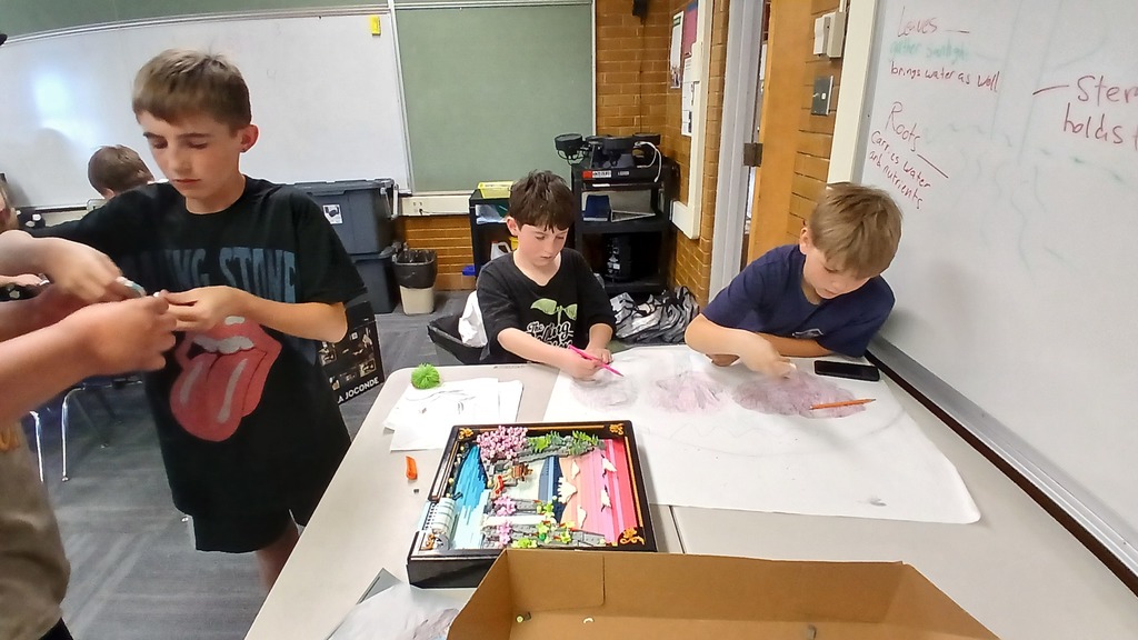 Three boys working at a classroom table. One boy is assembling Legos, while the other two work on a large drawing. A completed framed Lego art piece of a sunset over water sits on the table.