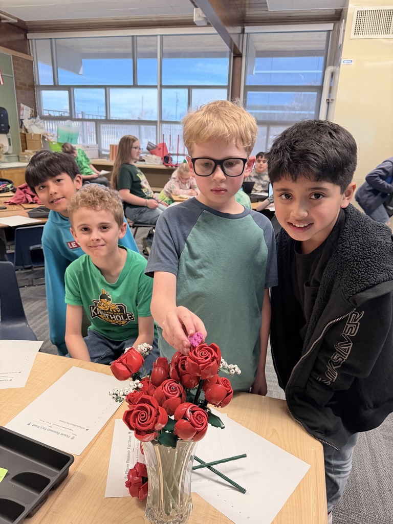 Four boys posing behind a desk featuring a realistic Lego botanical set of red roses in a glass vase. One boy is holding a small pink Lego heart.